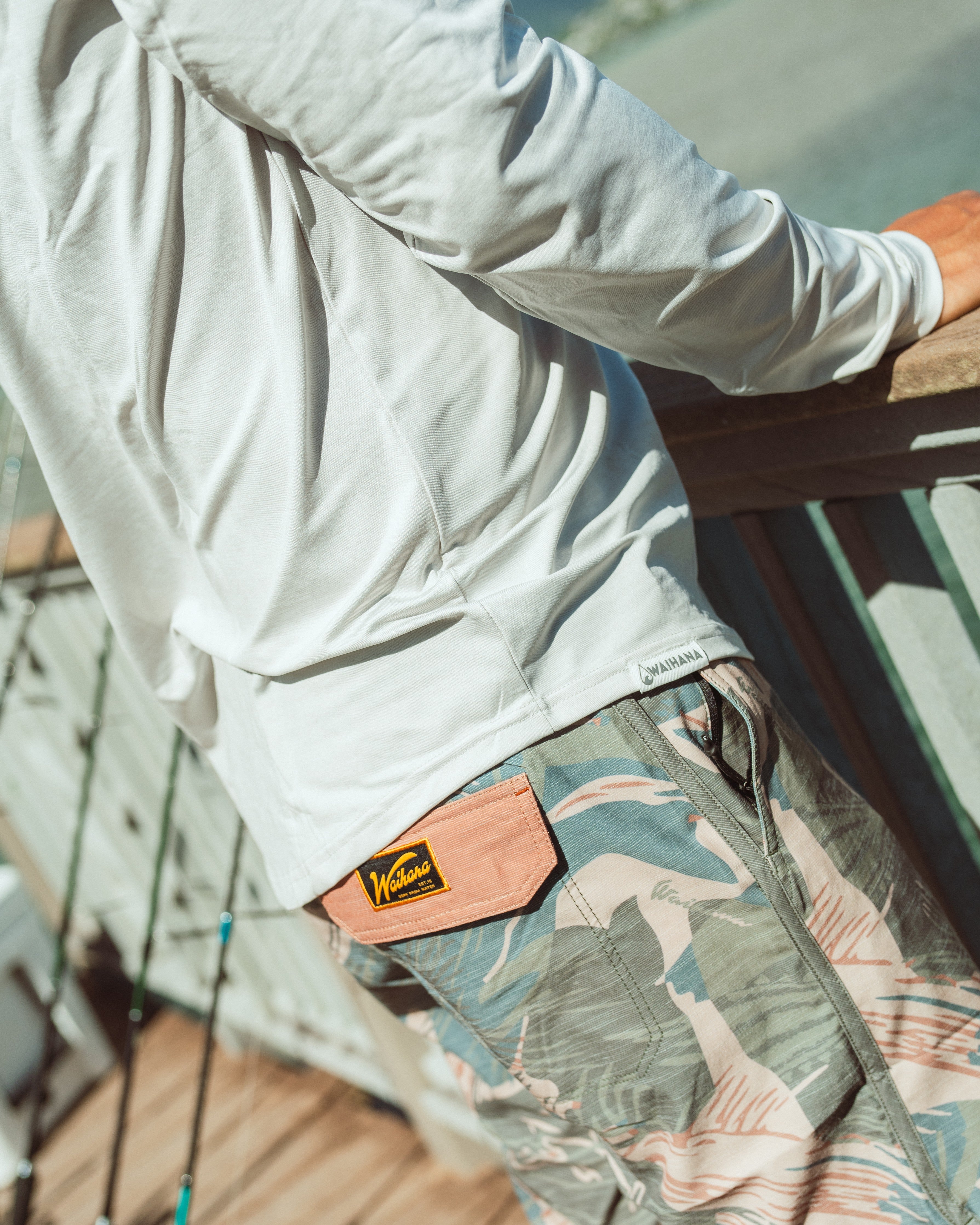 Close-up detail of Waihana Hawaiian Brushstroke Cruiser board shorts featuring a tropical leaf print with a coral-colored back pocket bearing the Waihana logo patch, worn with a white long-sleeve shirt on a wooden dock at Kaneohe Bay.