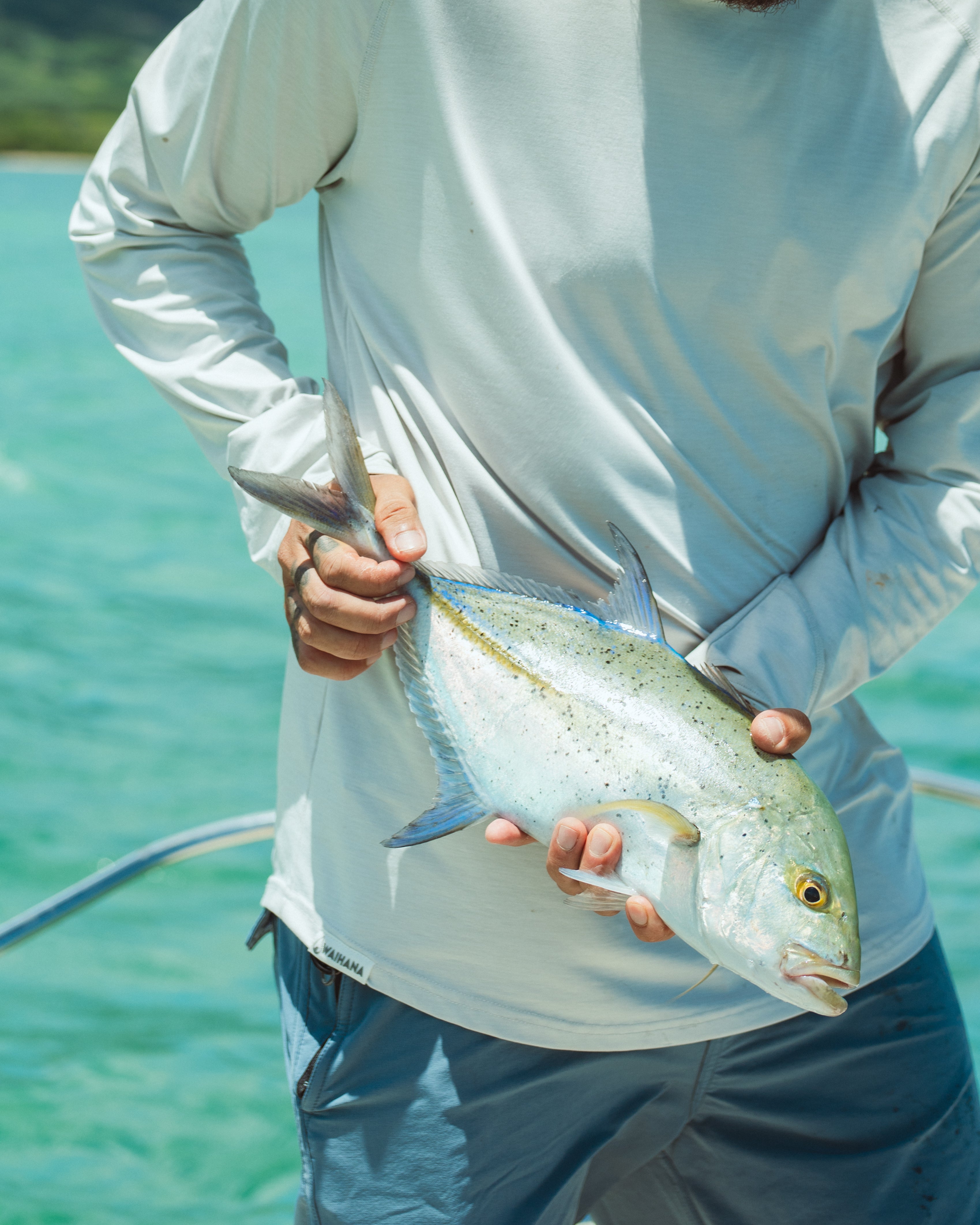 A man in a light grey Waihana long-sleeve rashguard holds a bluefin trevally fish with both hands against a backdrop of turquoise Kaneohe Bay water.